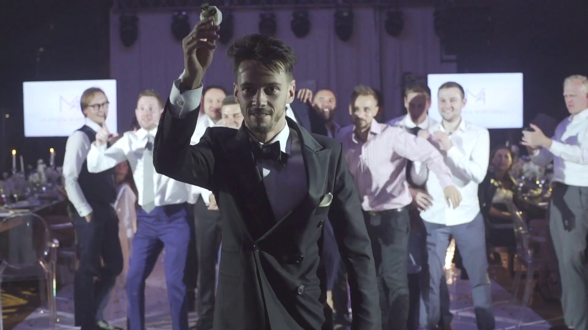 Man in a tuxedo raising a glass while guests cheer behind him at an evening celebration