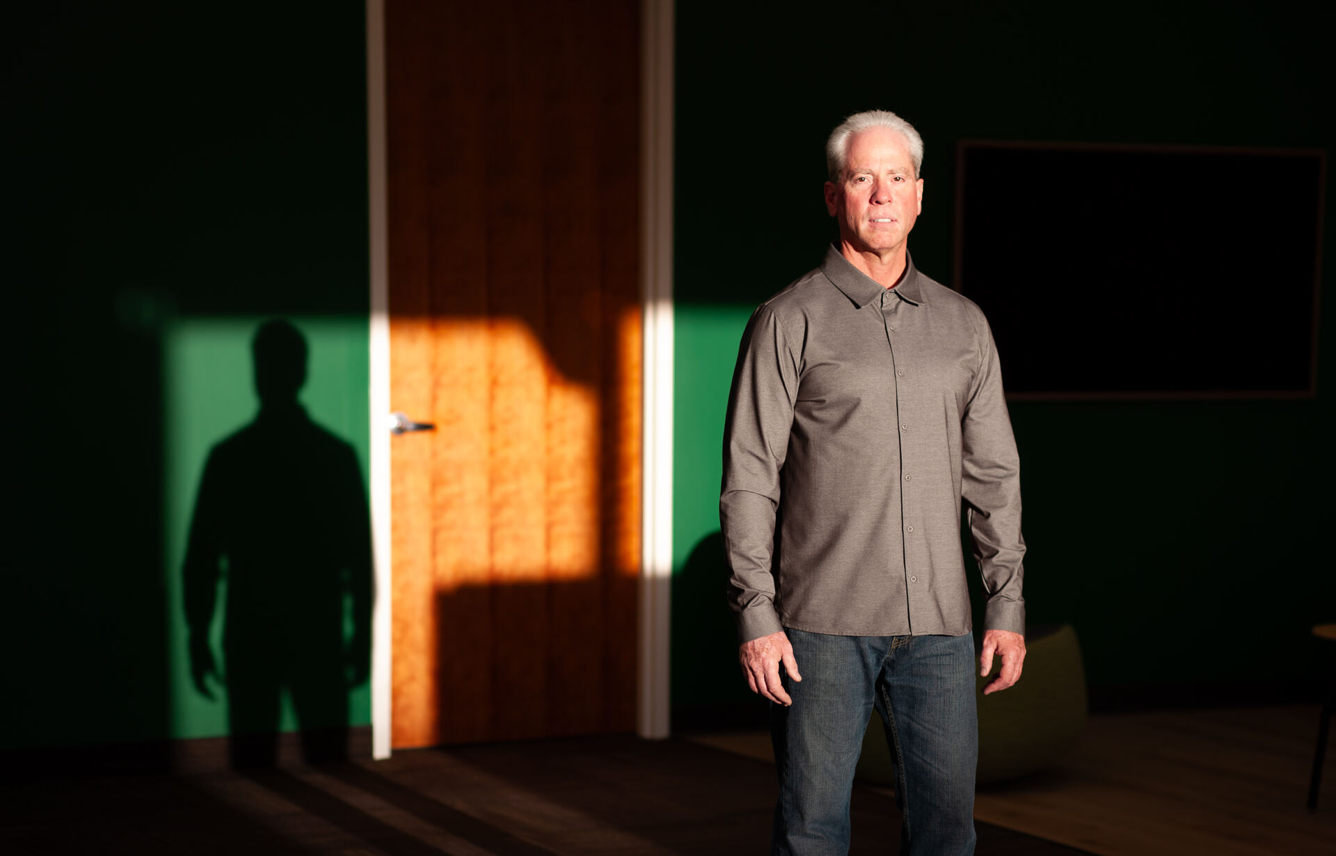 Man in a grey overshirt standing in a shaft of warm light against a dark green wall