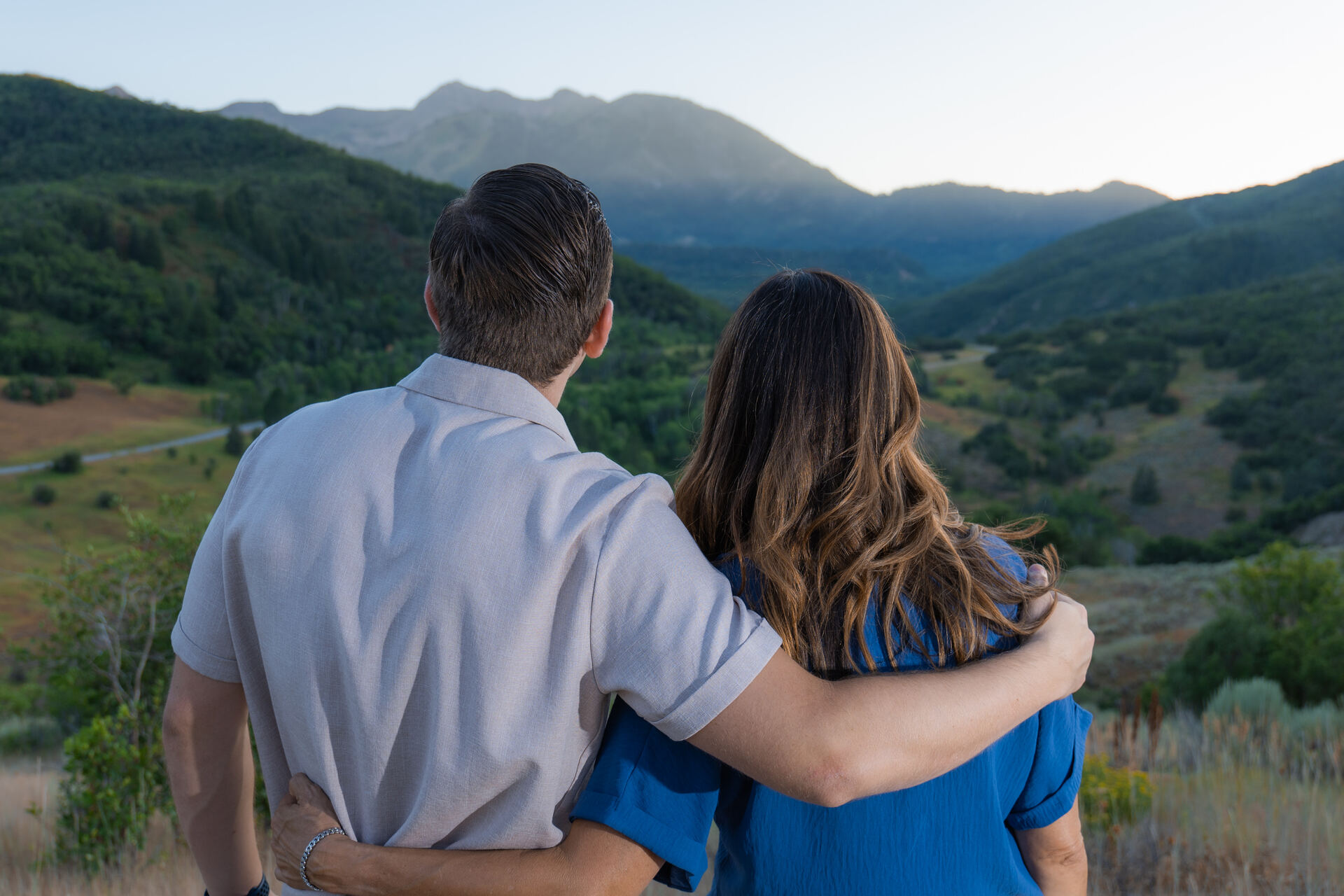 Couple seen from behind with their arms around each other, looking out over a green Utah mountain valley