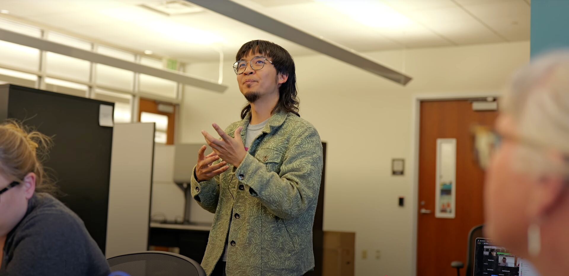 Man in a denim jacket speaking and gesturing in a sunlit office